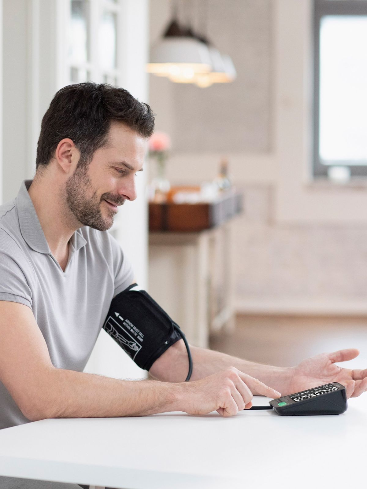 A man sits at a table and measures his blood pressure with a digital upper arm blood pressure monitor while pressing the start button.